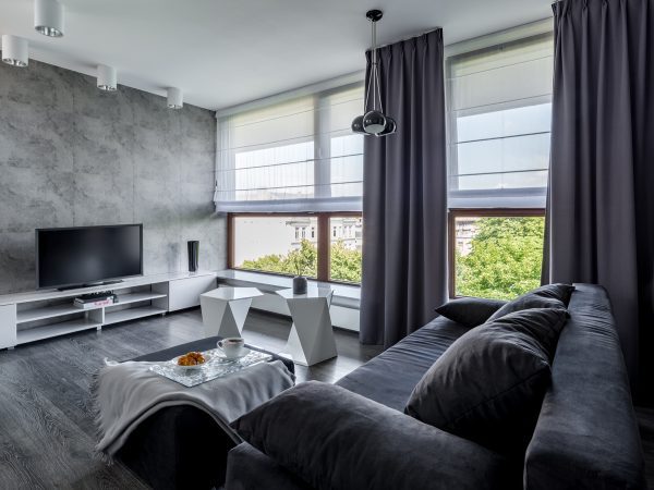 Modern living room featuring a dark sofa, TV on a stand, and an expansive window with gray curtains that reflect thoughtful window treatment pricing. The coffee table hosts a tray with a cup and snack, perfectly positioned to enjoy the serene view of trees outside.