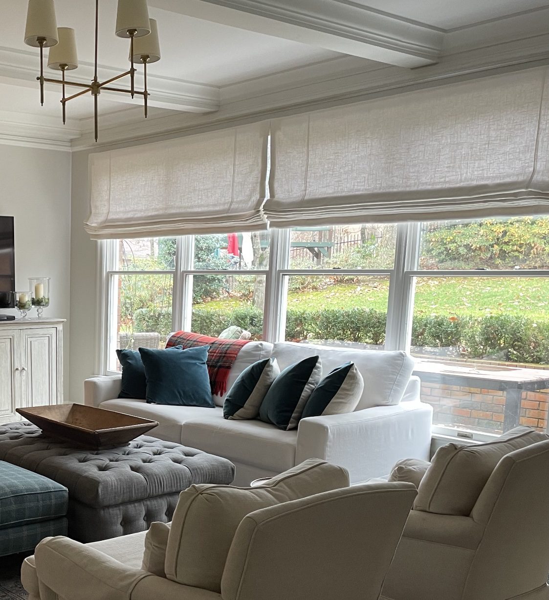 A living room with a white sofa, gray ottoman, and white armchairs showcases style and comfort. Multiple cushions and a plaid throw enhance the cozy vibe. Large windows feature sheer blinds, letting in natural light—a reason why Roman shades are so popular for their elegance and functionality.