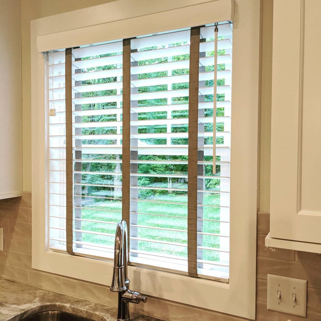 A kitchen window with white blinds, a silver pull cord, and a metallic faucet below showcases stylish kitchen window treatments, framing the greenery outside.