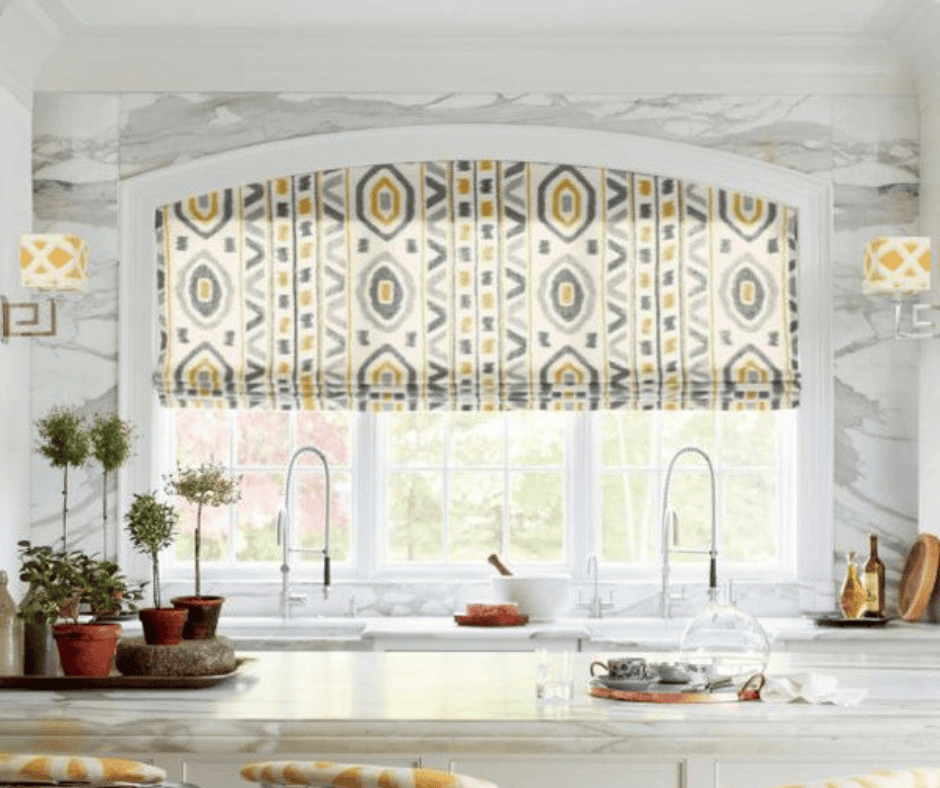 Kitchen with a marble countertop, two sinks, and a large window adorned with stylish kitchen window treatments like a patterned Roman shade. Potted plants and decorative items add charm to the space.