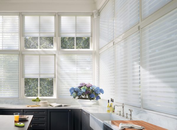 A kitchen with large windows featuring motorized white blinds and blue hydrangeas on the counter by the sink. Black cabinets, a wooden countertop, and a glass of orange juice are also visible.