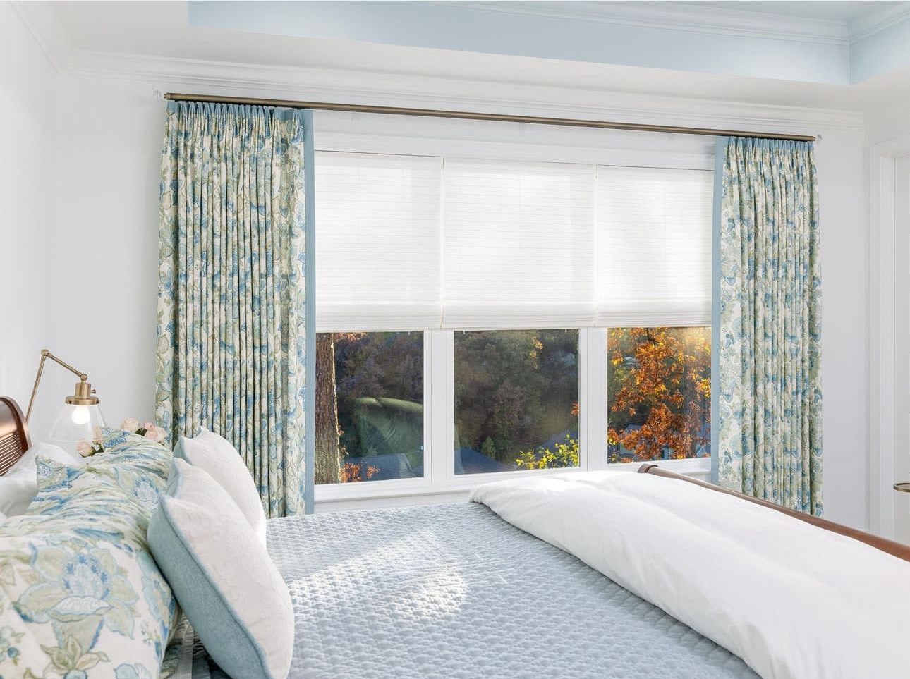 The bedroom features a large window adorned with blue and white floral curtains, perfectly complementing the blinds. The neatly made bed, dressed in a blue quilt and white pillows, adds to the room's calming color palette.