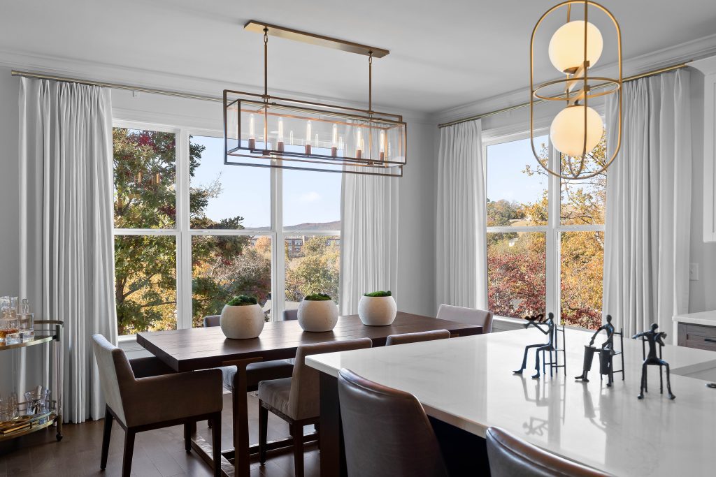 Modern dining room with a large custom wooden table, cushioned chairs, decorative plants, and a view of trees outside. Contains a bar cart, pendant lighting, and small figurines on a marble counter—a testament to why custom is worth the investment in style and functionality.