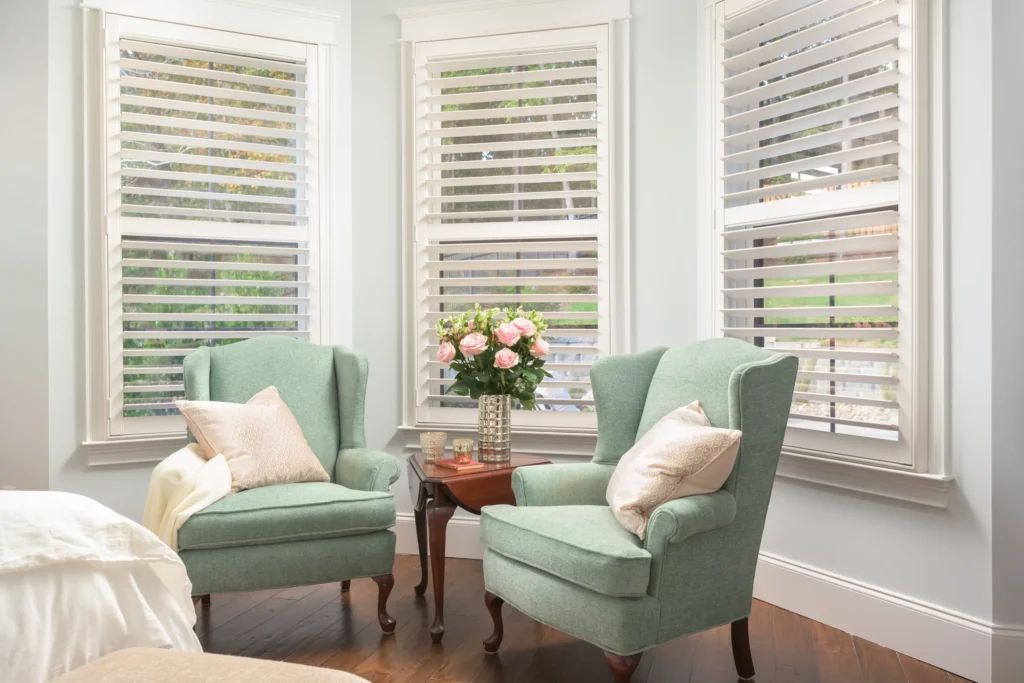 Two green armchairs with cushions are placed around a small wooden table with a vase of pink flowers. The background features three windows with white shutters.