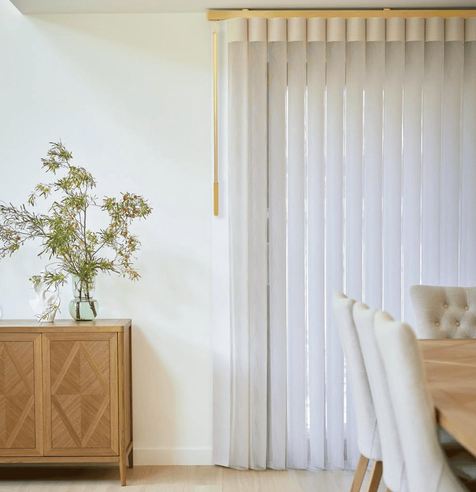 A dining room with a wooden table, upholstered chairs, a sideboard with a vase of greenery, and vertical white blinds covering a large window.