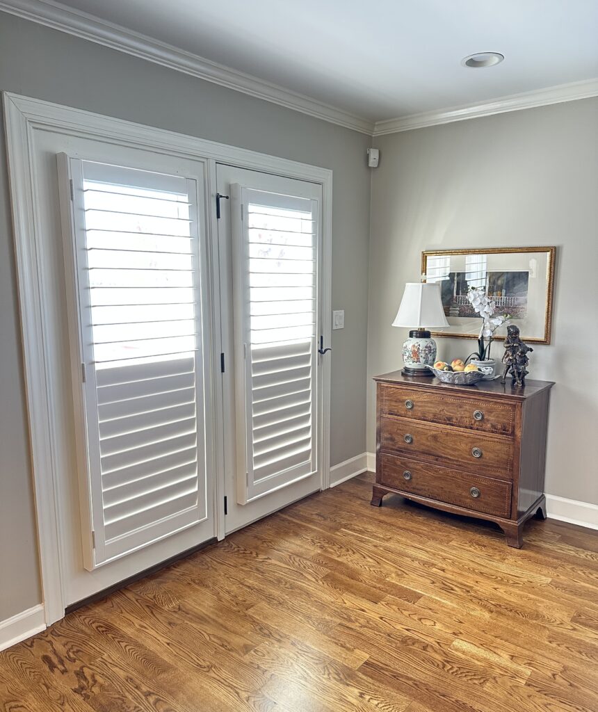 A pair of white shuttered patio doors next to a wooden chest of drawers with a lamp, decorative items, and a framed picture above, all on a hardwood floor.