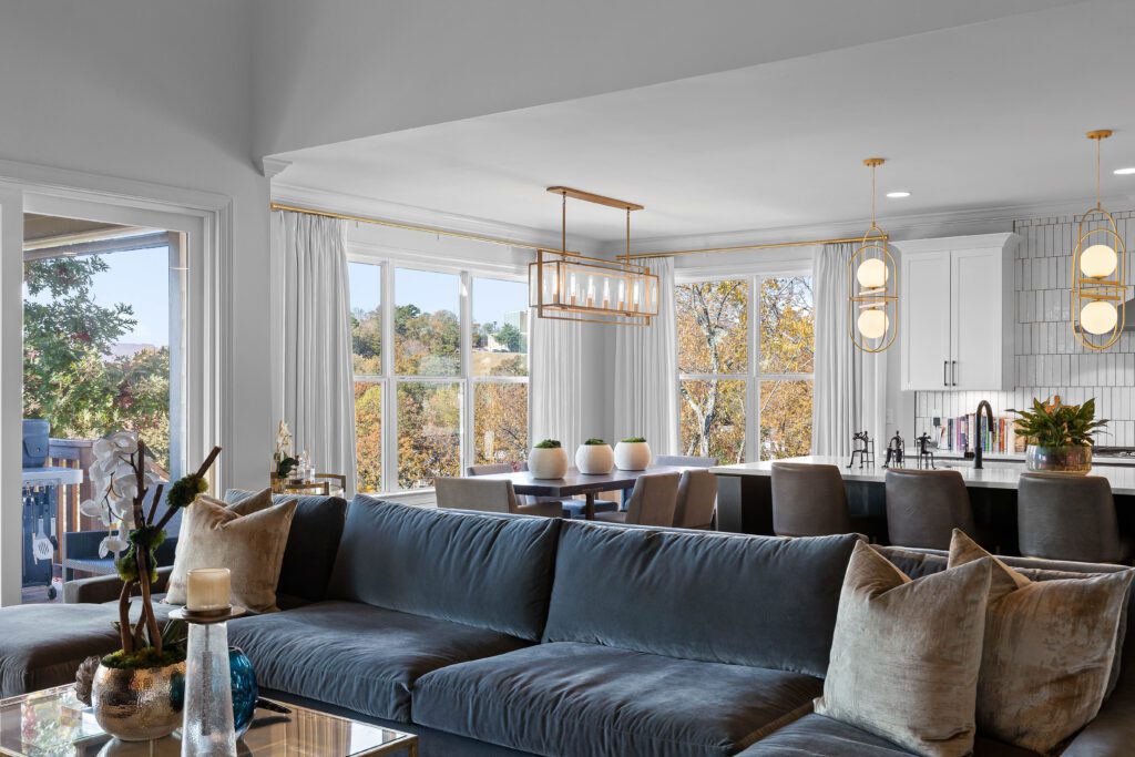 Modern open-concept living room and kitchen with large windows, gray sectional sofa, dining table, and pendant lighting. Trees and outdoor scenery visible through the windows.