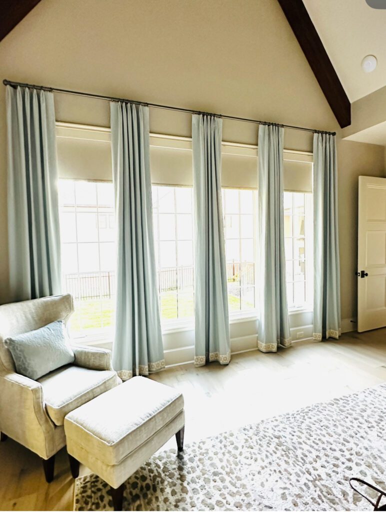 A bright living room features large windows with pale blue curtains, an armchair with an ottoman, and a light-colored patterned rug.