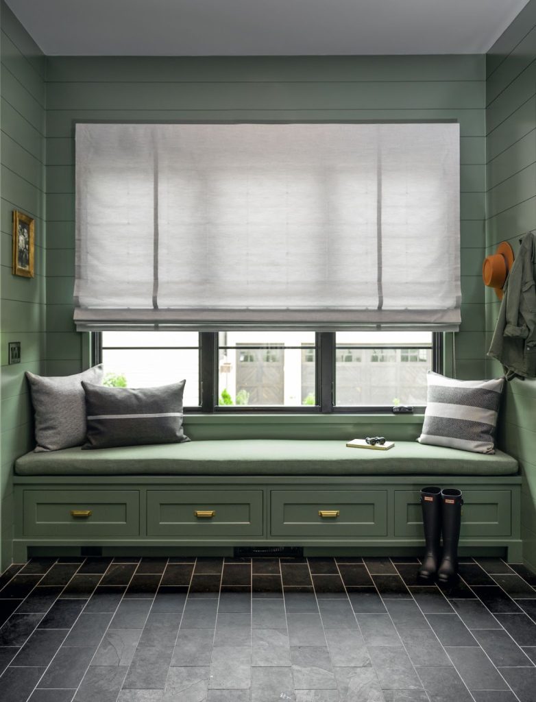 A room features a green cushioned window seat crafted with Sunbrella fabric, pillows, drawers beneath, white blinds, and slate flooring. A coat and hat hang on the wall. Rubber boots are on the floor.